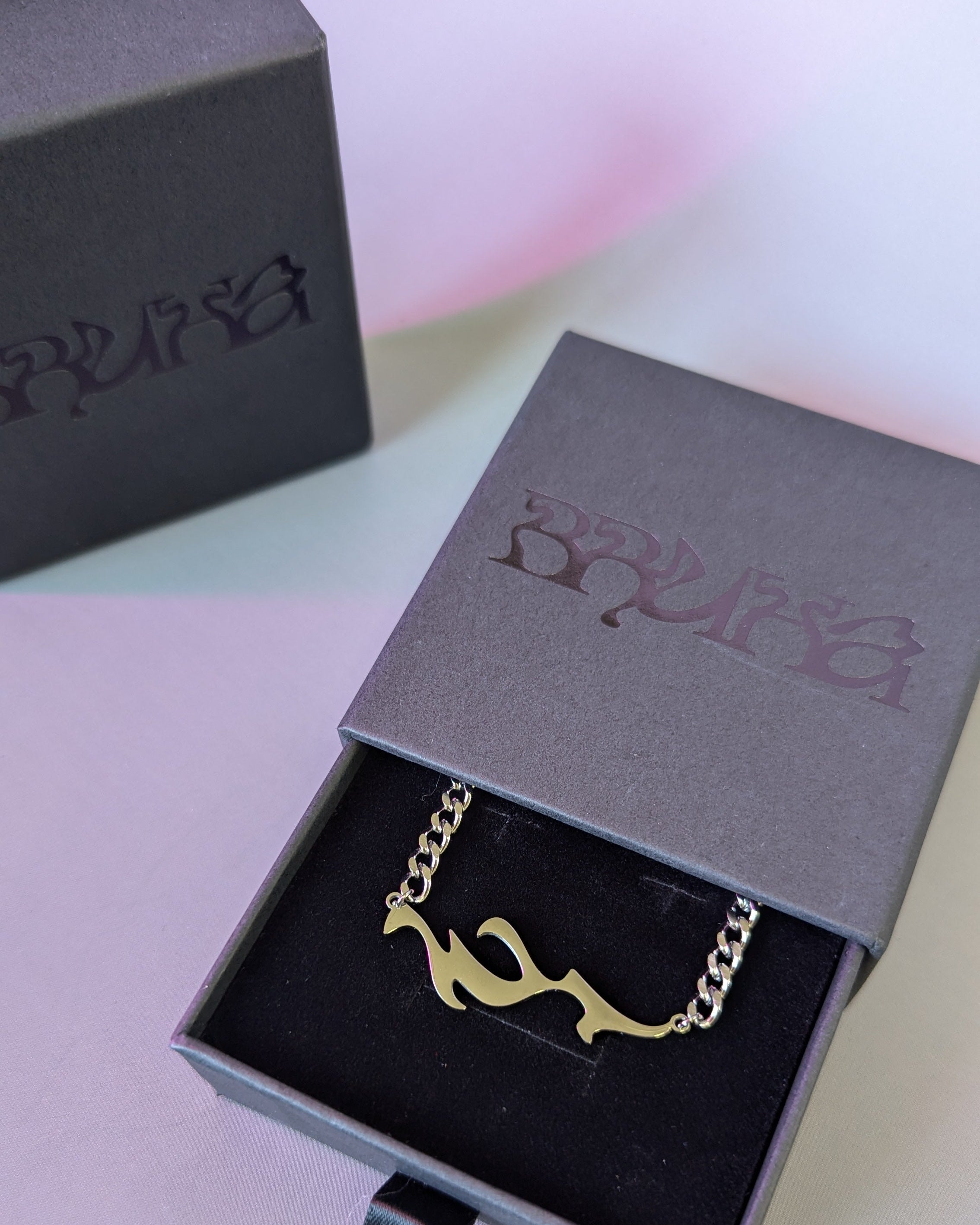 Gold necklace in an open gray jewelry box with a visible brand logo on a light pink background.